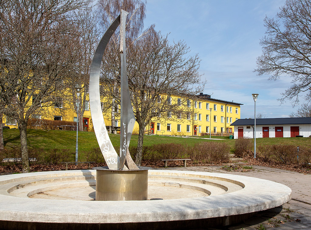 Metallskulptur i en rund fontän, gula hus bakom.
