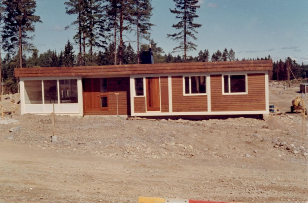 Enplansvilla i mörkt trä med vita knutar på grusplan.