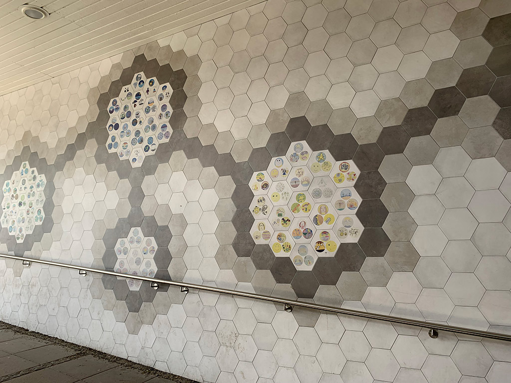 19 sexkantiga kakelplattor med små bilder på, omgivna av grått kakel. Också det med sex kanter. Flera fält med kakel som sammanfogas med linjer av gråa kakelplattor.