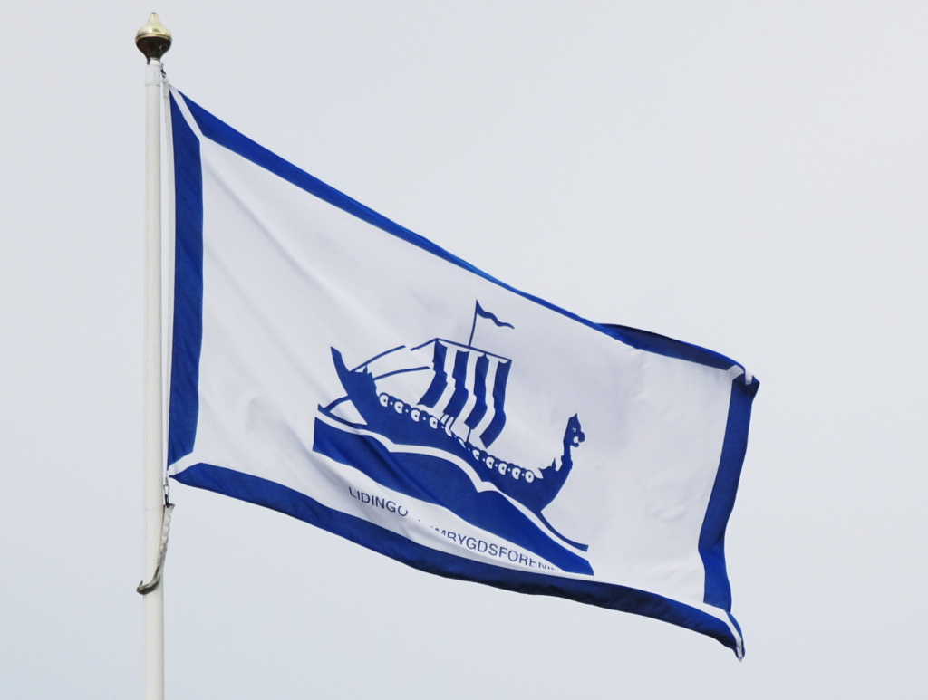 Flagga med Lidingö stads kommunvapen. Flaggan är blå och vit med ett stiliserat vikingaskepp på.