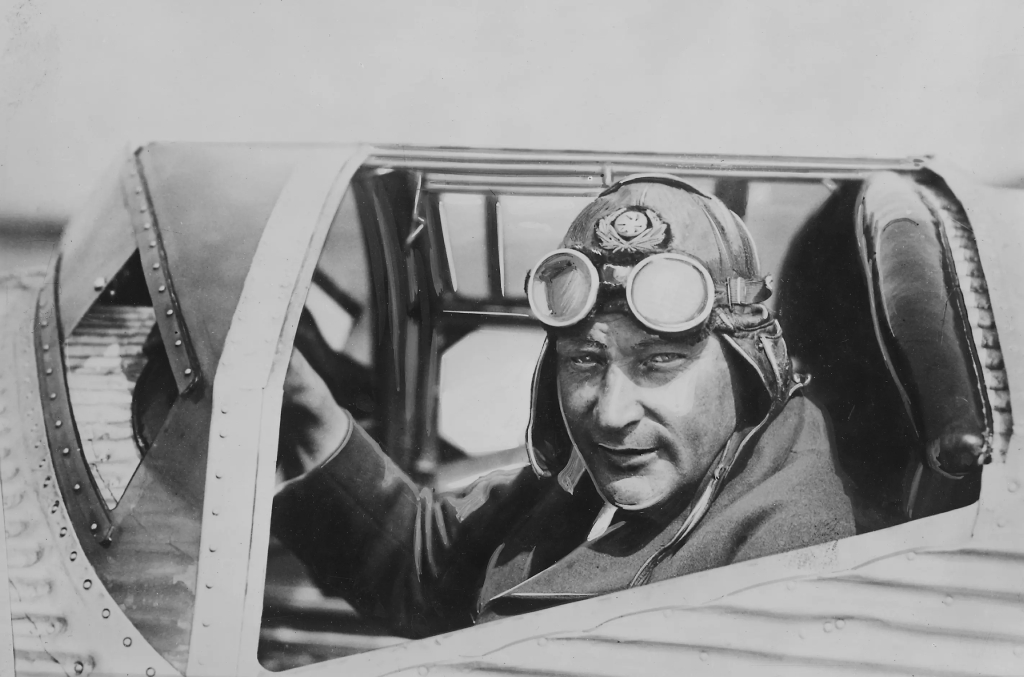 Närbild av en man som sitter i en flygplanshytt med skinnmössa och flygarglasögon.