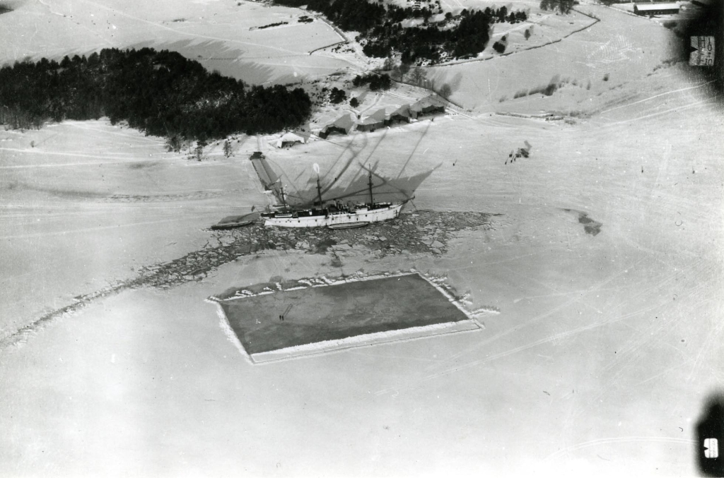 Ett flygfoto med stranden i ovankant, is på vattnet, en plogad rektangel på isen och ett uppbruten is runt det vita fartyget.