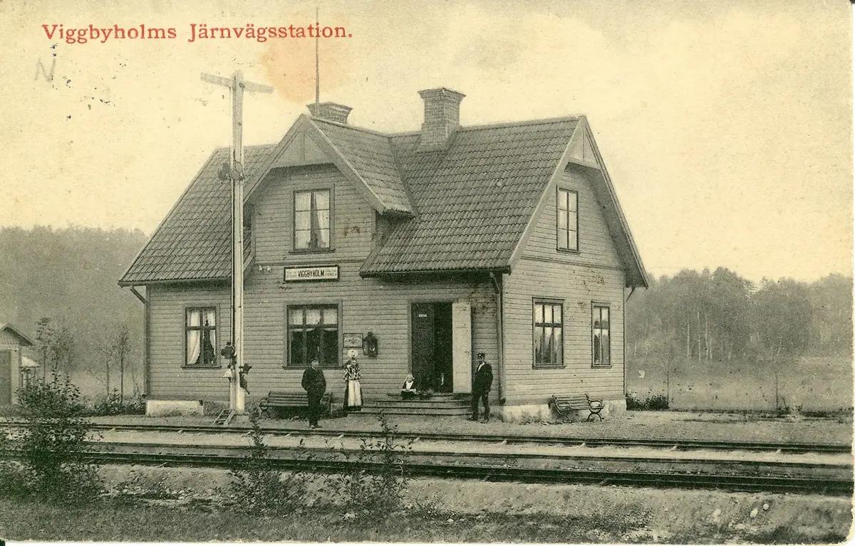 Ett svartvitt foto av stationshuset. Två järnvägsspår i förgrunden. Två män och en kvinna står framför huset och en liten flicka sitter på trappan.