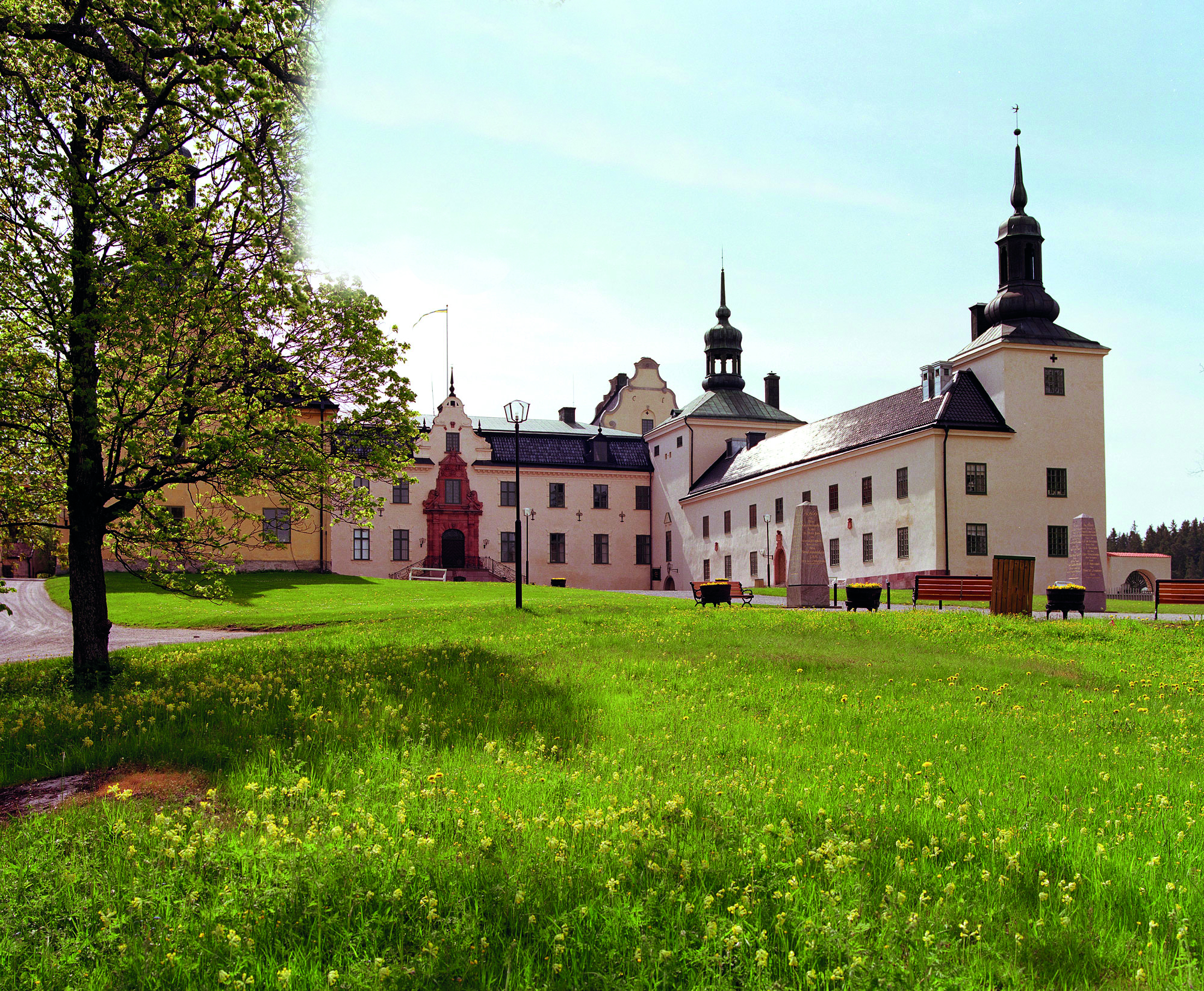 Slott bakom en stor gräsmatta med gula blommor. I mitten syns slottets monumentala ingång med trapp och stor dörr.