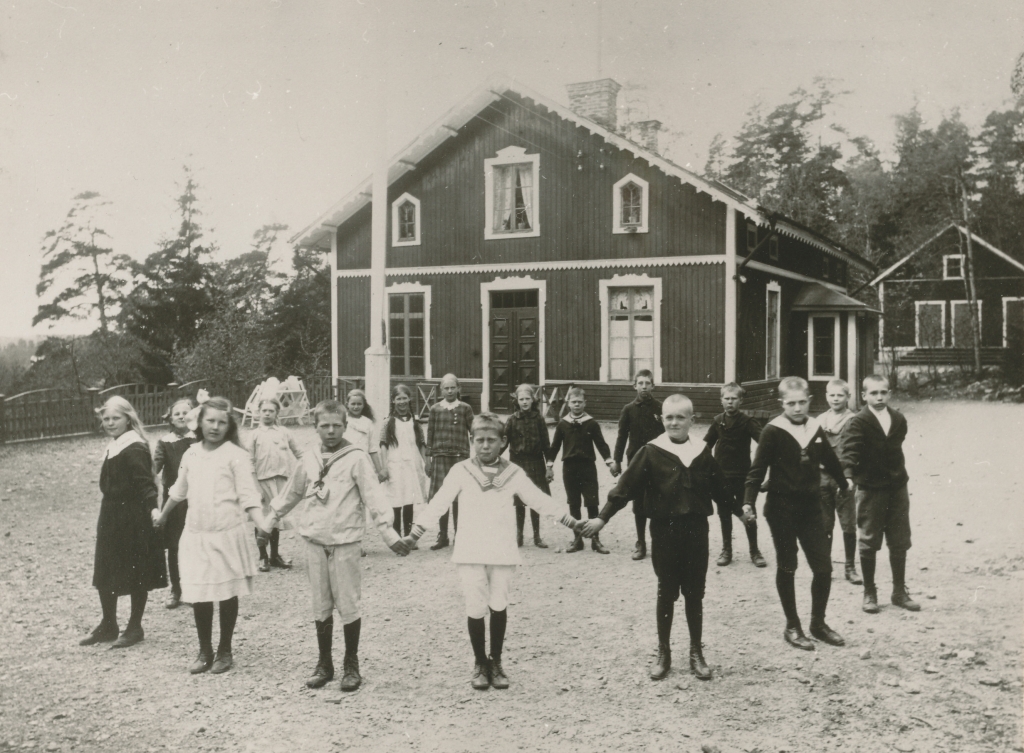 En skolklass står i ring och håller varandra i händerna. Skolbyggnaden i trä och två våningar syns i bakgrunden.