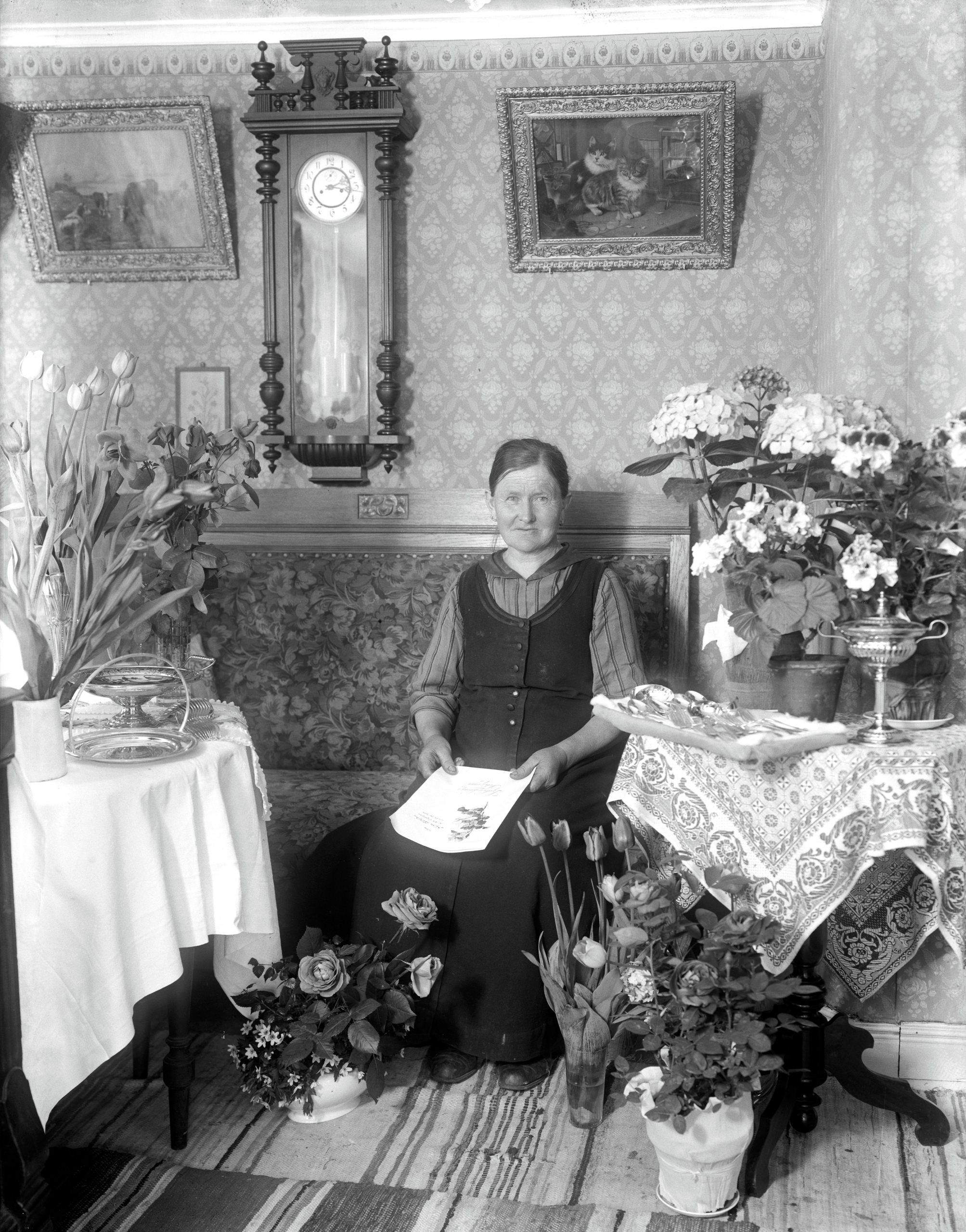 Svartvitt foto där Mina sitter i en salong omgiven av blombuketter. Trasmattor på golvet, medaljongtapet, väggur och tavlor på väggen.