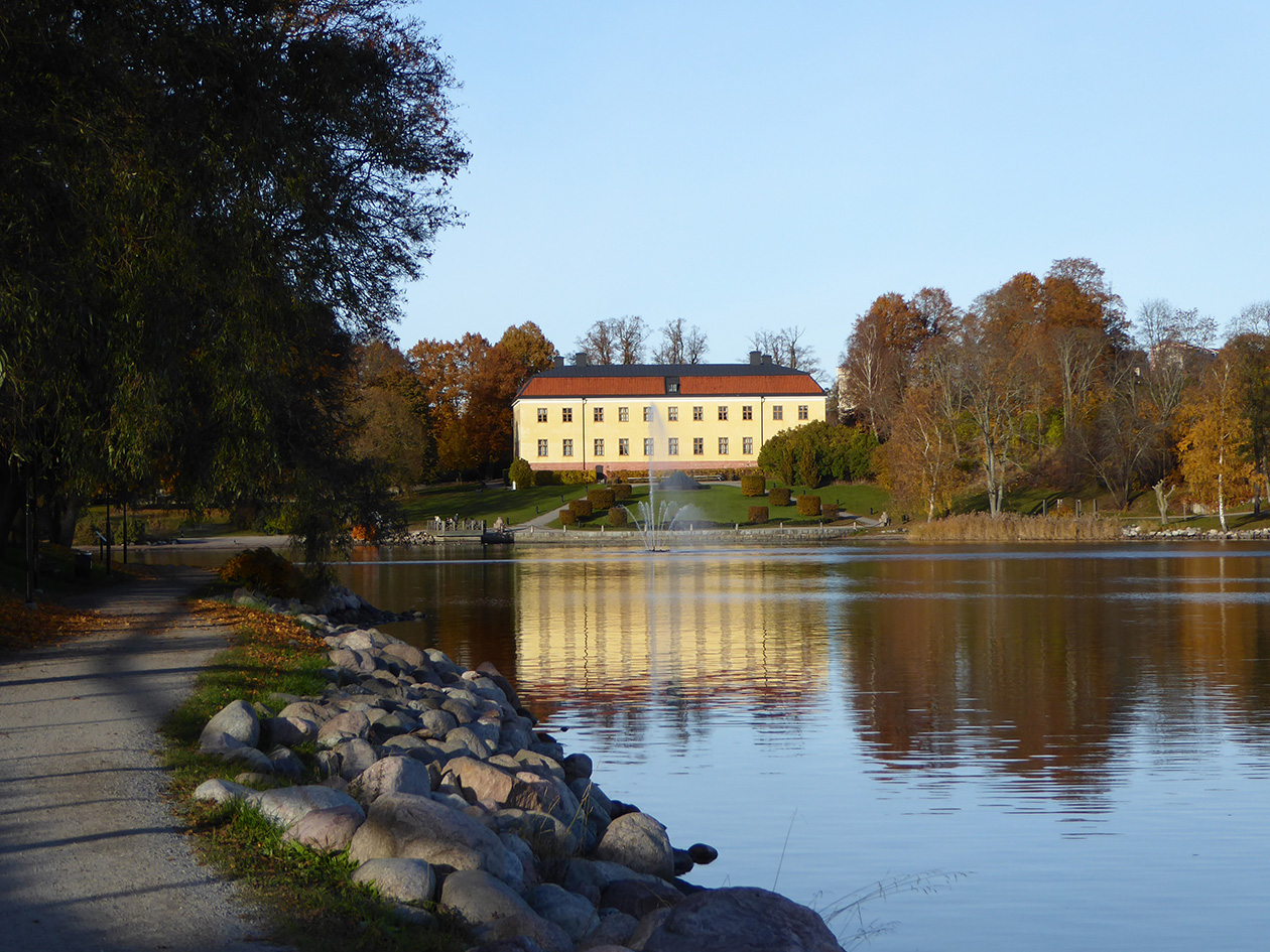 Ett vitt stort stenhus speglar sig i en sjö. I förgrunden en stensatt strandkant.