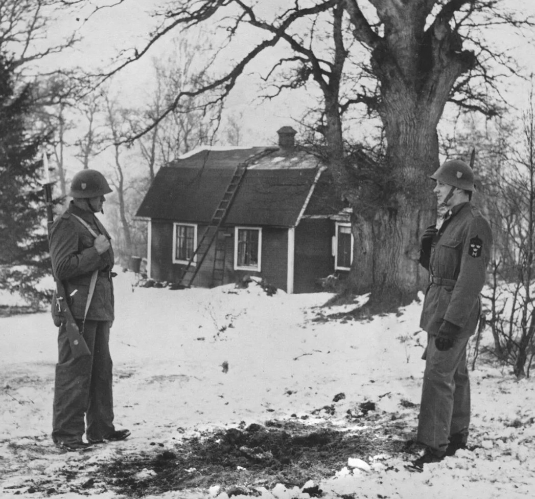 Svartvitt foto där två militärer står på var sin sida av en svart fläck i den vita snön, med stugan i bakgrunden.