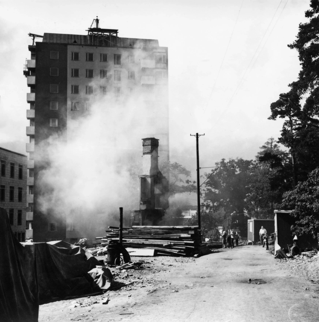 En skorsten från ett nedbränt hus sticker fram ur röken. Ett höghus i bakgrunden.