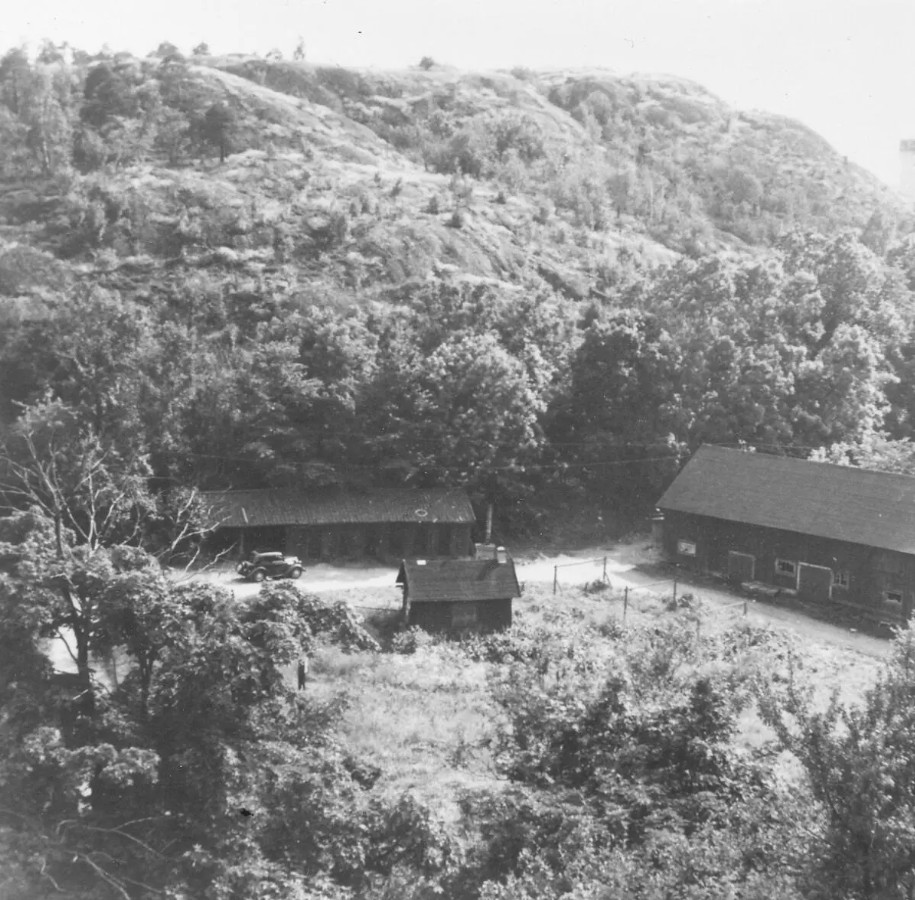 Svartvitt foto där fotografen blickar ner mot tre enkla trähus, omgivna av grönska. En äldre bil syns också och ett högt obebyggt berg i bakgrunden.