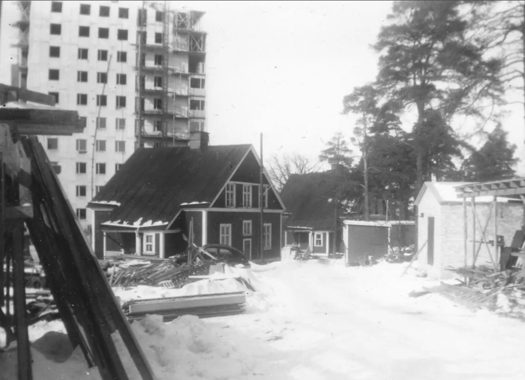 Svartvit bild där ett högt punkthus under byggande sticker upp bakom två äldre trähus. vinter.