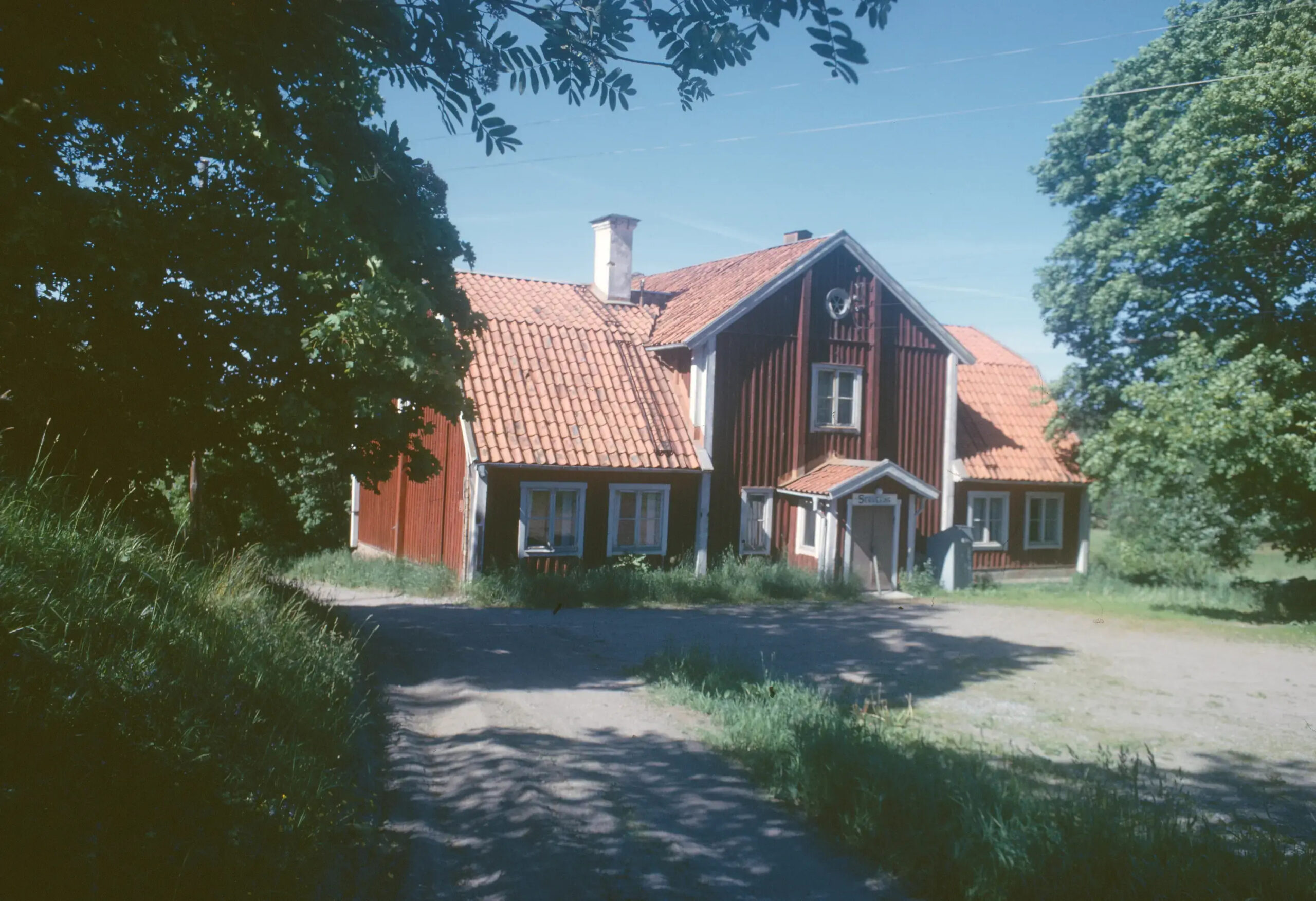 En färgbild som visar det röda trähuset med vita gavlar och en ganska tom gårdsplan en sommardag.
