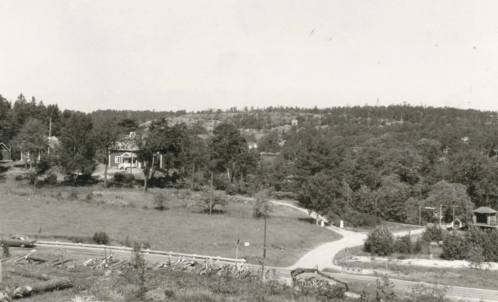 En bil kör på en smal landsväg. Bortom den syns en järnvägsövergång, ett par hus på en kulle. Vattnet skymtar bakom de grönskande träden och på andra sidan syns kala klippor och skog.