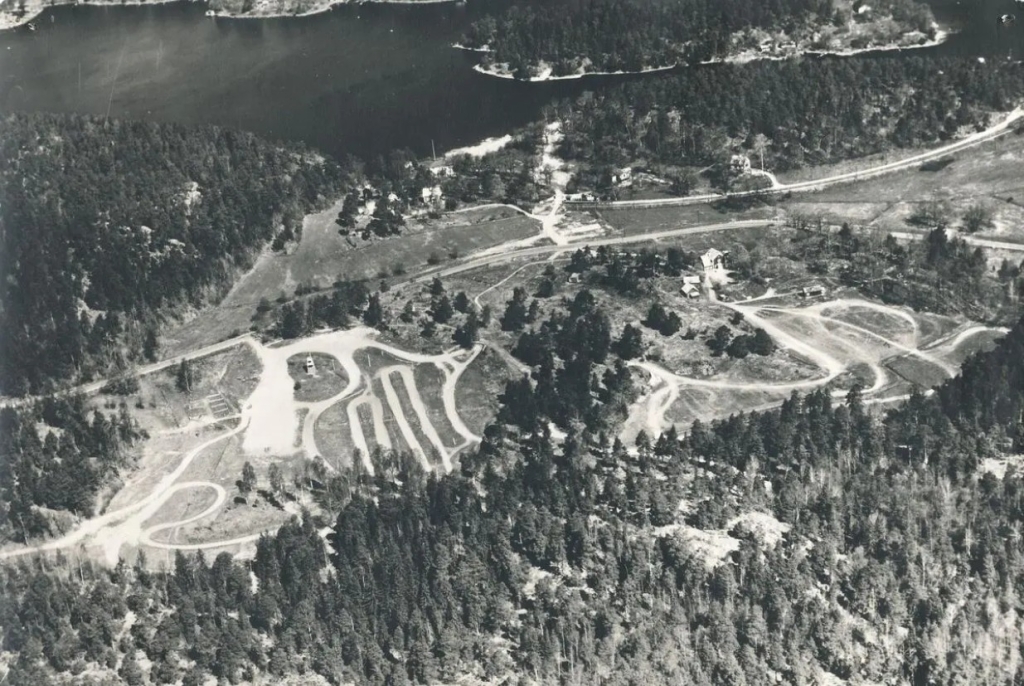 Flygfoto som visar några utspridda hus, järnvägen, en bilväg och motocrossbanan.