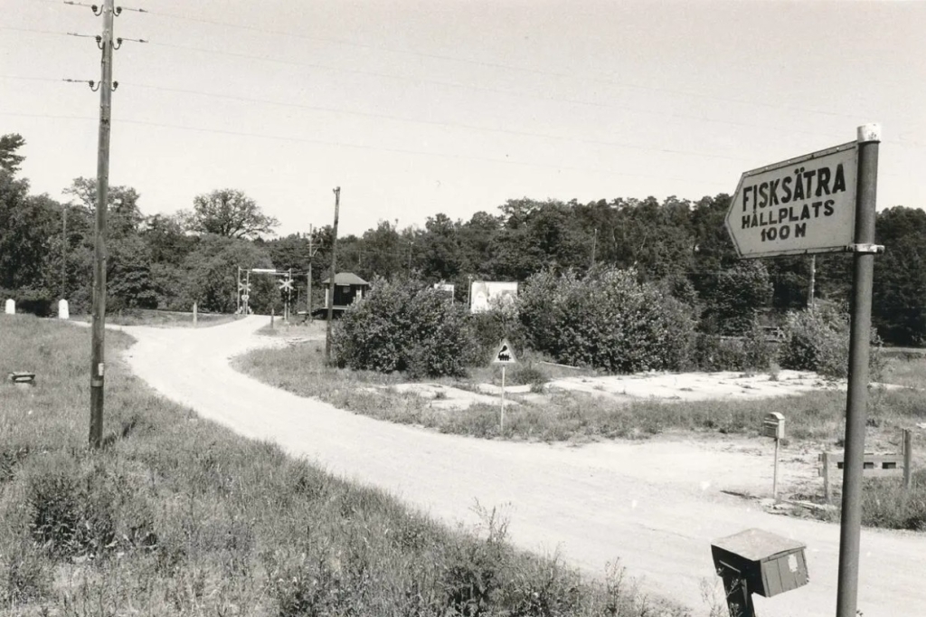 Svartvit foto med en skylt i förgrunden som säger "Fisksätra hållplats 100 meter" en grusväg leder fram till en järnvägsövergång med en liten väntekur.