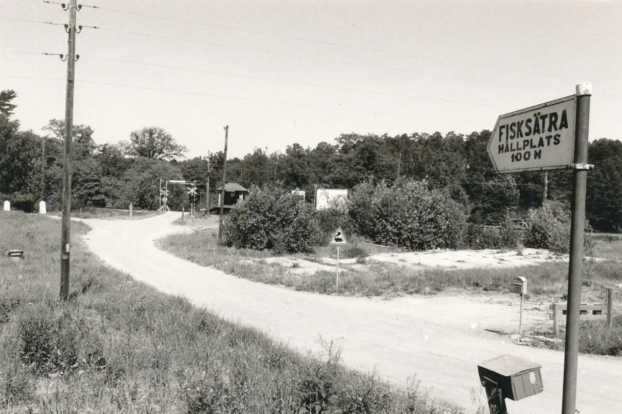 Svartvit foto med en skylt i förgrunden som säger "Fisksätra hållplats 100 meter" en grusväg leder fram till en järnvägsövergång med en liten väntekur.
