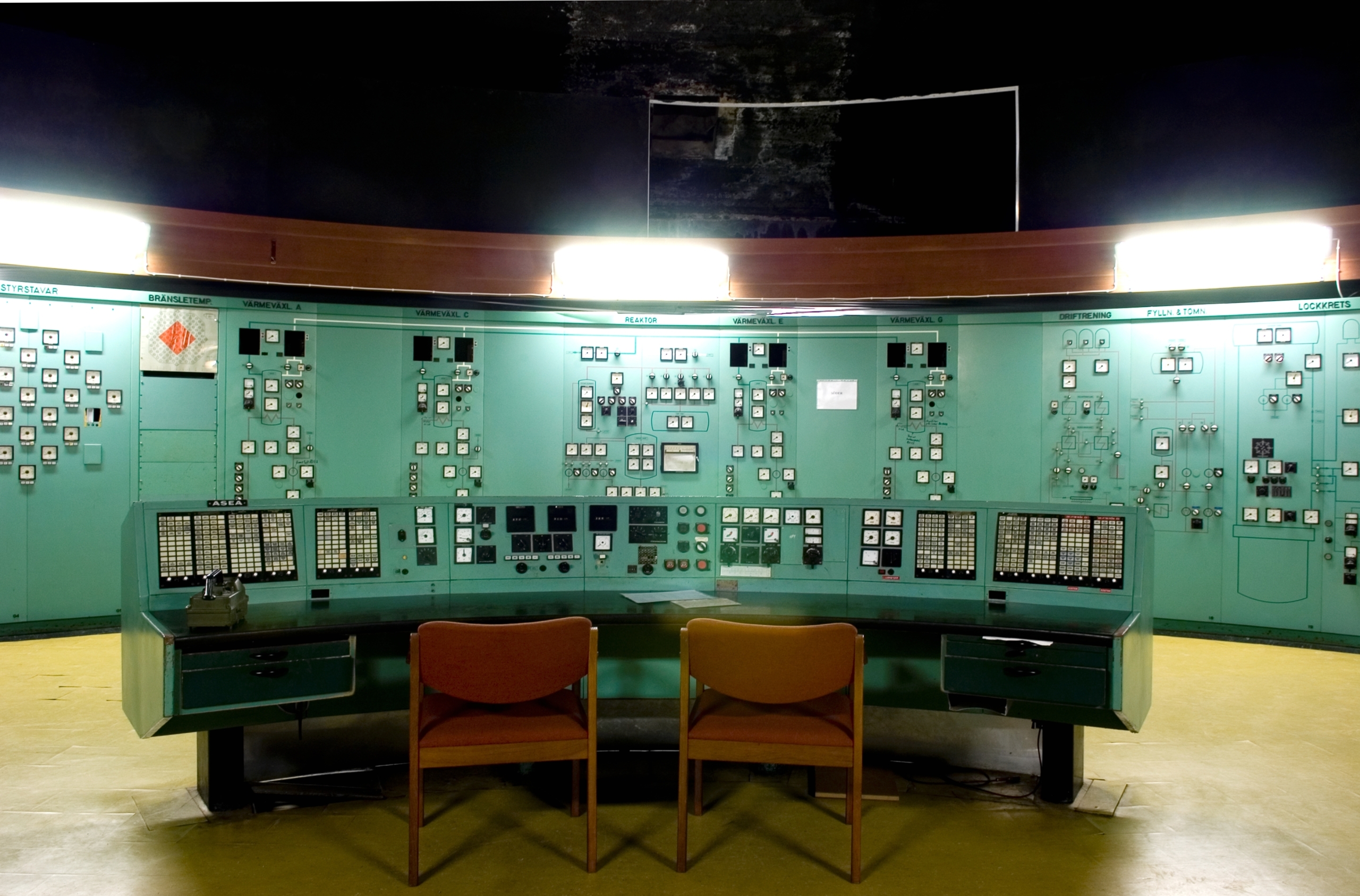 Kontrollrum med gröna paneler, lysramp i trä och två trästolar vid kontrollbordet.
