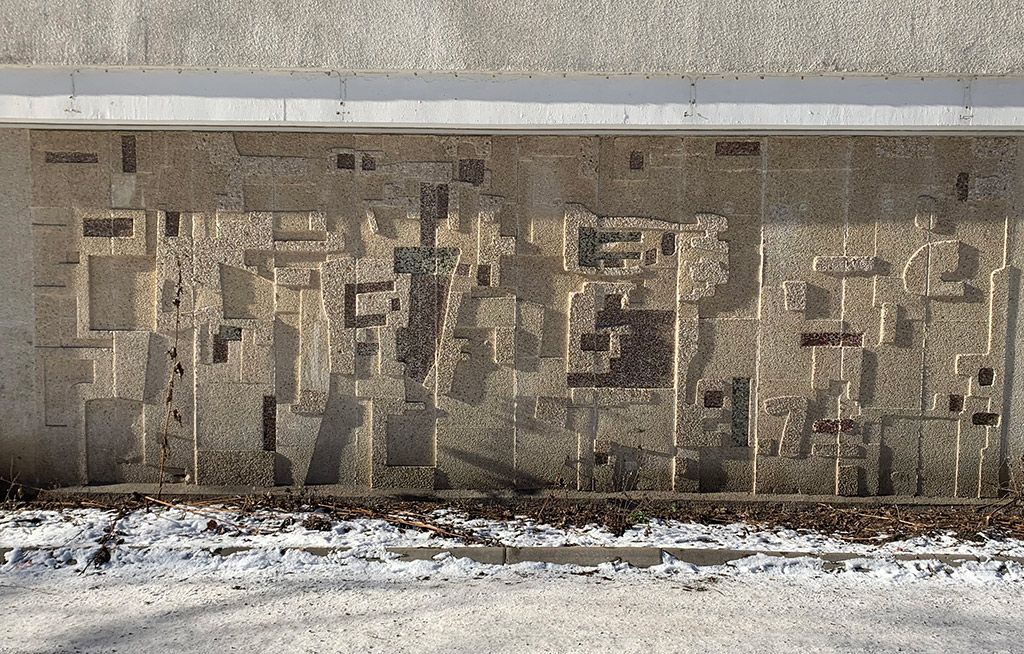 Abstrakt konstverk i relief på vägg. Grundfärgen är beige med inslag av grönt och brunt.