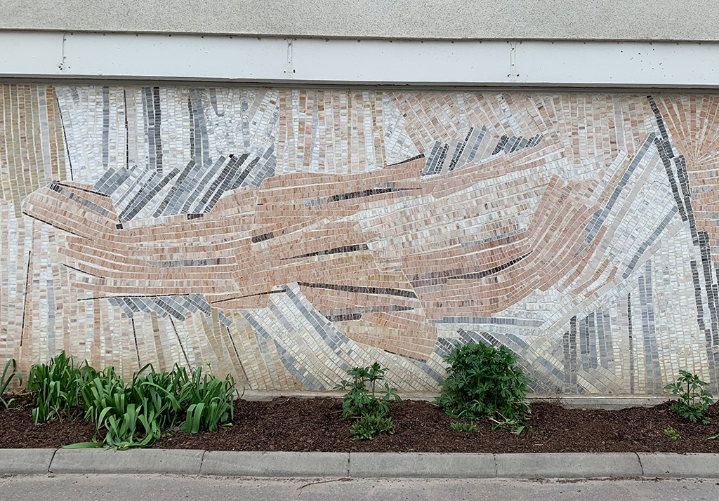 Mosaik på vägg. Ett fantasidjur i ljusa bruna nyanser med inslag av grått.