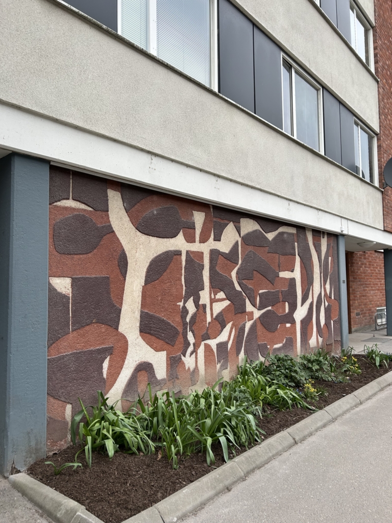 Abstrakt konstverk på sockeln på ett hus. Konstverket är gjort i bruna, beiga färger med former som går in i varandra.