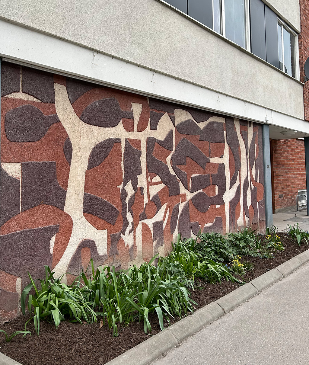 Abstrakt konstverk på sockeln på ett hus. Konstverket är gjort i bruna, beiga färger med former som går in i varandra.