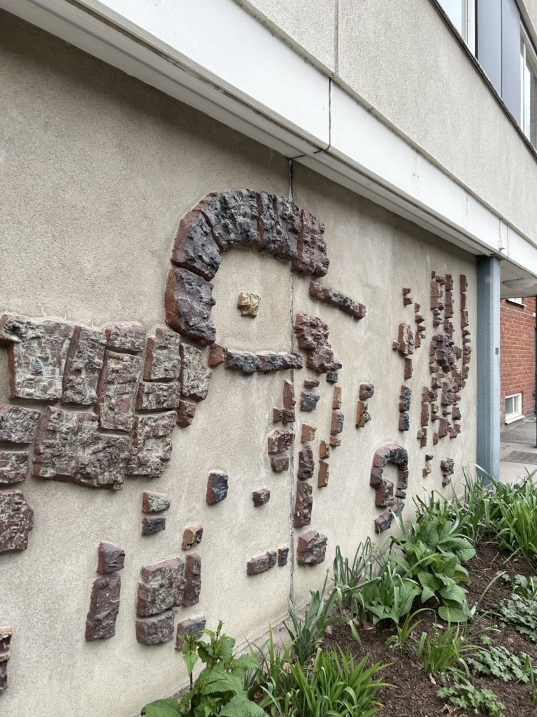 Abstrakt konstverk med utspridda betongbitar i guldfärg och tegelfärg på en betongsockel på ett hus. Rabatt med gröna blad framför.