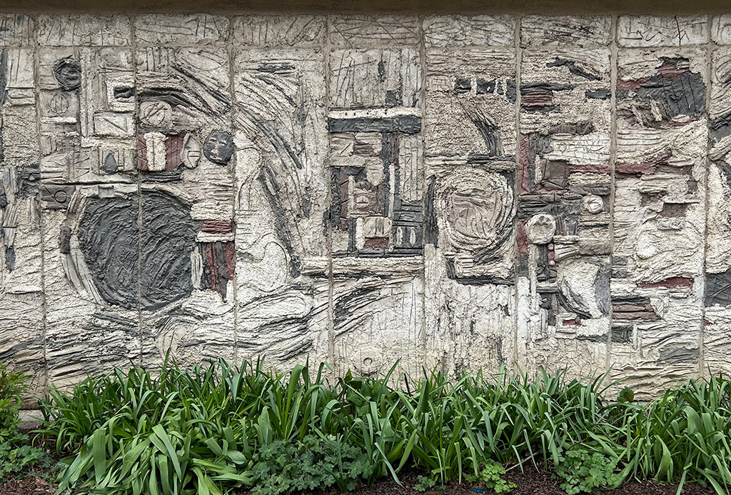 Abstrakt verk av grå betong, och med inslag av mörkt grått och brunt. Betongen är lagd i relief med ristningar i. Rabatt med gröna växter framför.