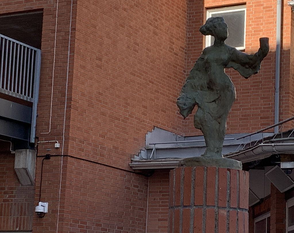 Skulptur av kvinna i brons gjord i abstrakta former. Står framför en röd tegelbyggnad.