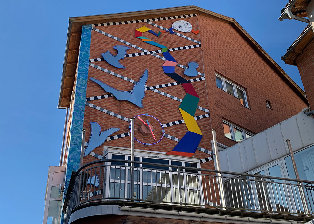 Abstrakt konst i på en vägg på en byggnad av tegel. Ett sicksackband i svart och vit löper över hela väggen. Fem ljusa former är utspridda. Högst upp en figur och från den vecklar ett band ut sig i färgerna rött, gult, blått, rosa, svart, grönt och orange.