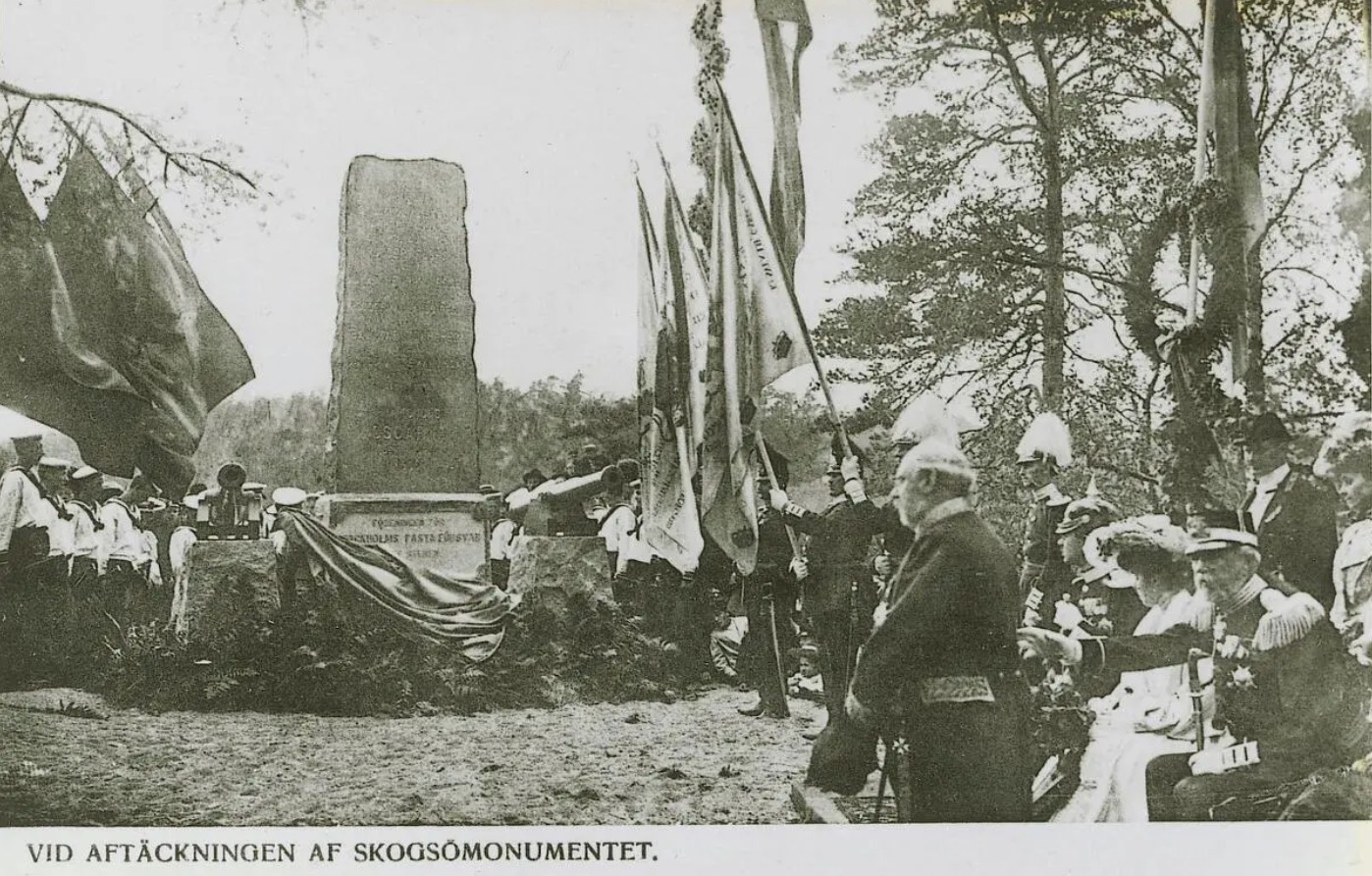 Täckningen faller från stenmonumentet. Militärer med flaggor och kransar. Kungaparet sittande.