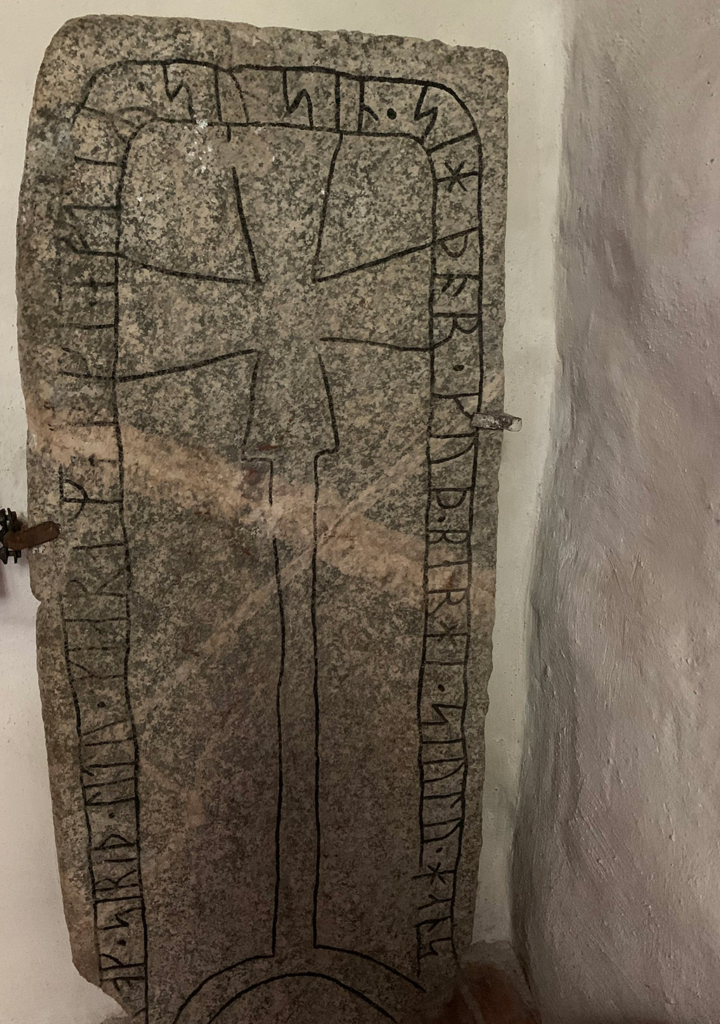 Runsten med kors i mitten och runor runtomkring. Inomhus med vit vägg bakom.