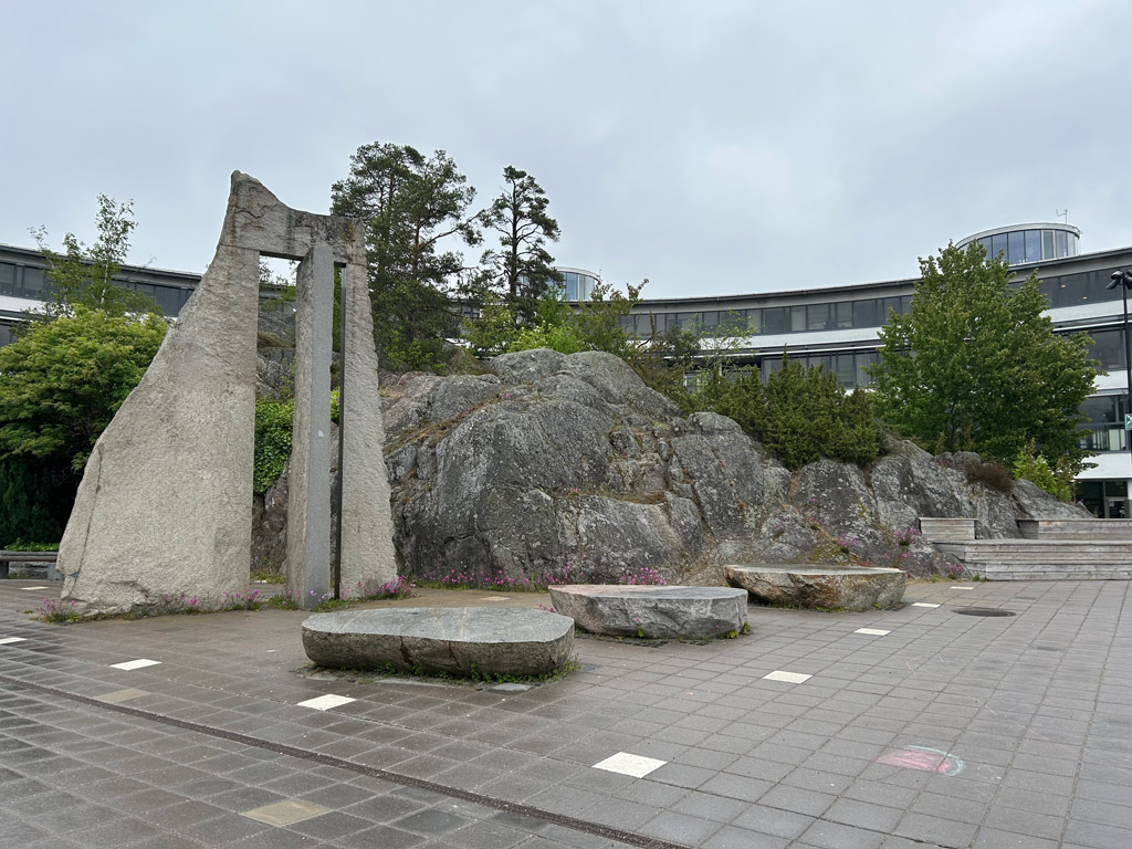 Ett torg med en hög stenskulptur och en klippa bakom. Framför är tre släta slipade stenar i låg höjd.