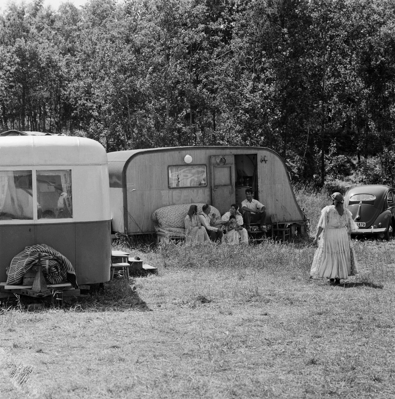 Några vuxna och ett par barn sitter utanför några äldre husvagnar och en bil. En Kvinna i klänning går mot personerna