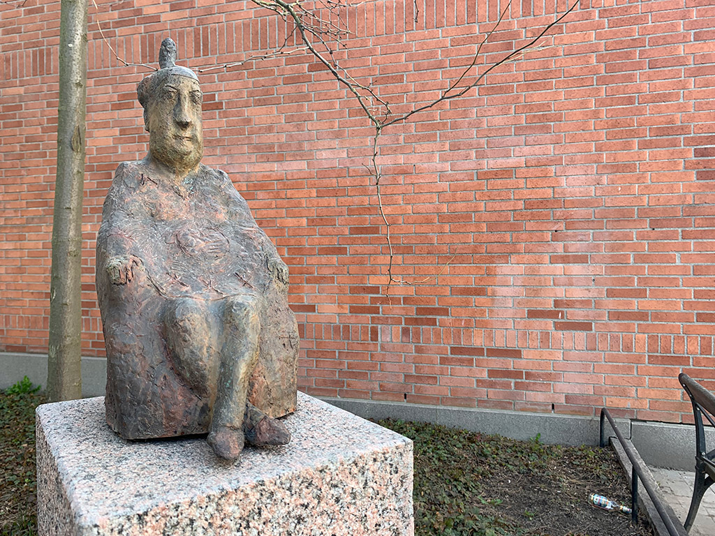 Skulptur av en sittande kvinna med benen i kors och med en smal uppstickande form på huvudet.