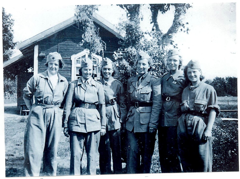 Sex kvinnor uppställda i uniformer framför ett hus på landet.