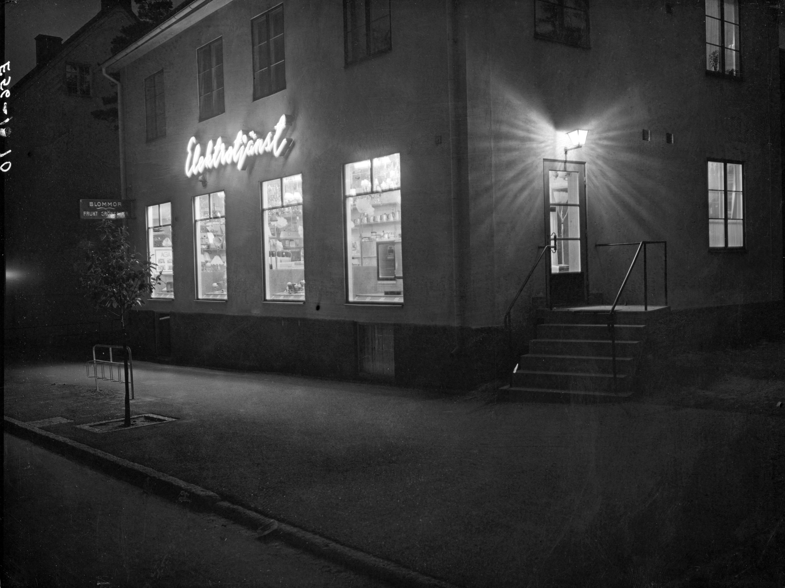 Ljuset flödar från fönster, dörr och neonskylt med texten Elektrotjänst. Ut på den mörka gatan.