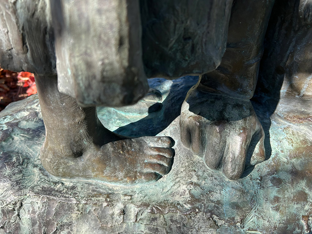 Detalj av en skulptur i brons. En barnfot och en knoge som lutar mot underlaget av mindre obearbetad brons.
