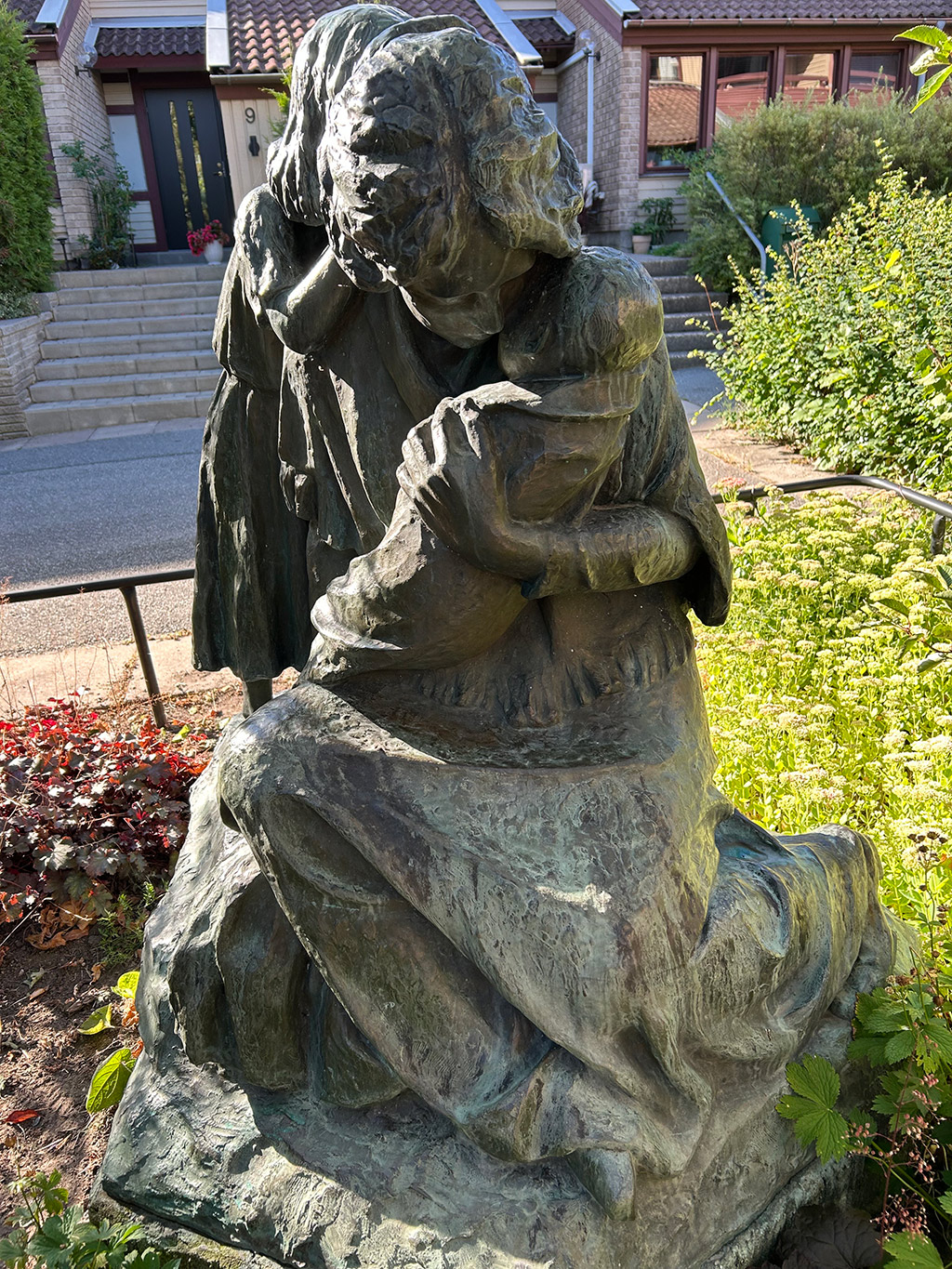 Skulpturgrupp med en kvinna som håller en bebis och en flicka som står bakom kvinnan. I en park med grönska.