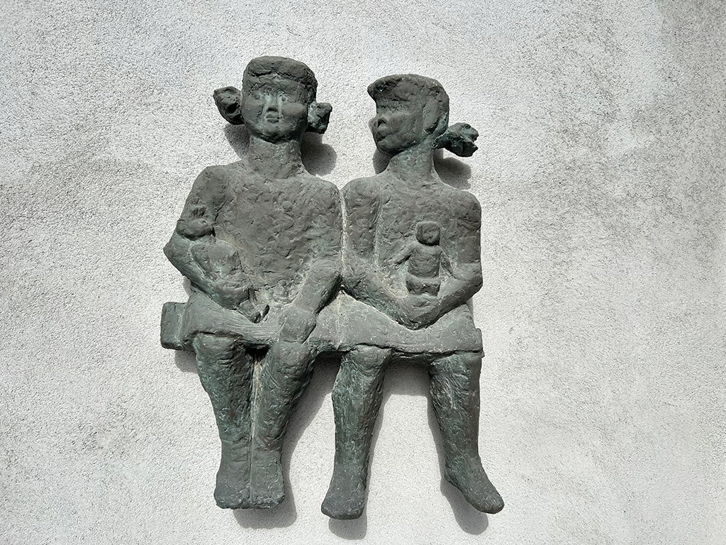 Bronsrelief på grå vägg med två flickor som sitter med dockor i famnen.