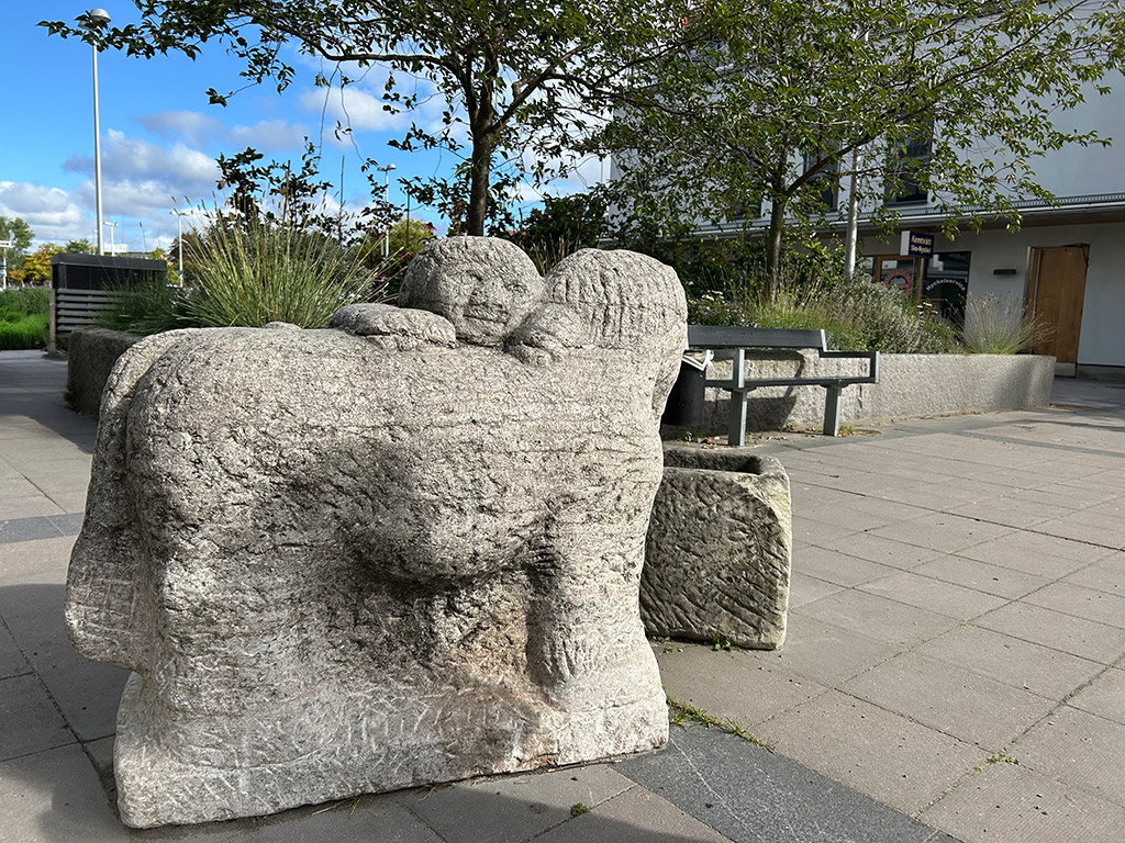 Skulptur av en häst med runda former i sten med ho framför. Torgmiljö.
