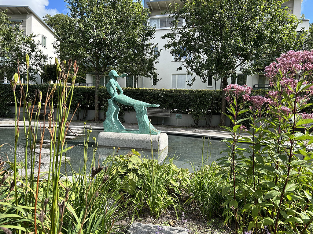 En grön skulptur av en sjöjungfru i en damm. Blommor och grön växtlighet längst fram. Till höger rosa höga blommor.