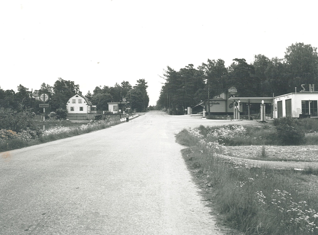 Svartvitt foto. Shellmack, konsum och en villa kantar den asfalterade vägen utan målade streck.