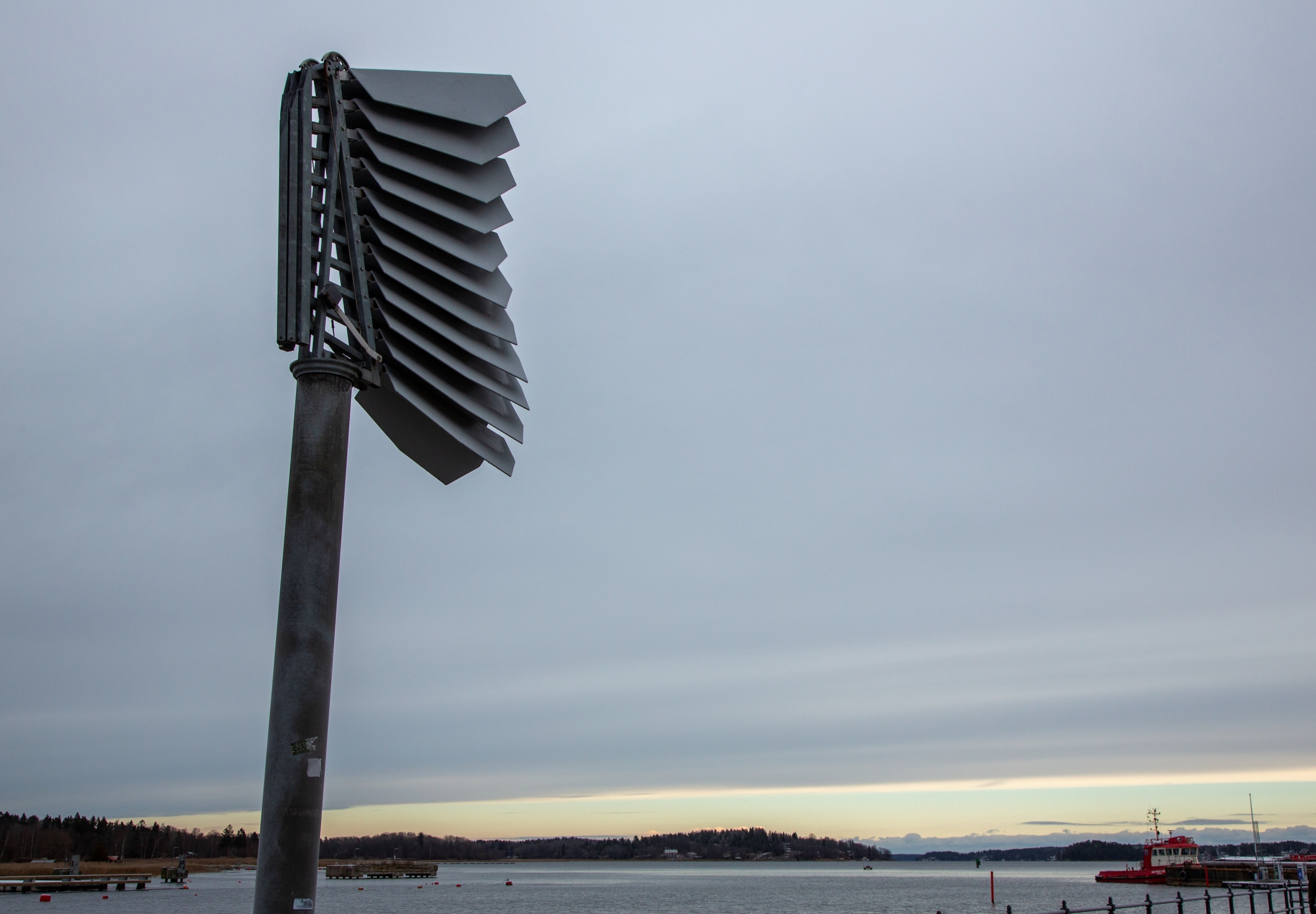 Hög skulptur av stål som vidgar och veckar sig i. På en pelare. Himmel och vatten bakom.
