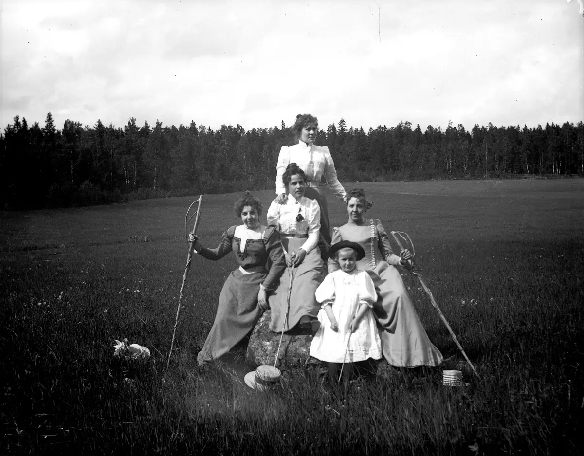 Svartvitt foto från sekelskiftet 1900. Fyra kvinnor och en ung flicka står i grupp på en äng. Flera av dem håller i långa spetsiga träkäppar.